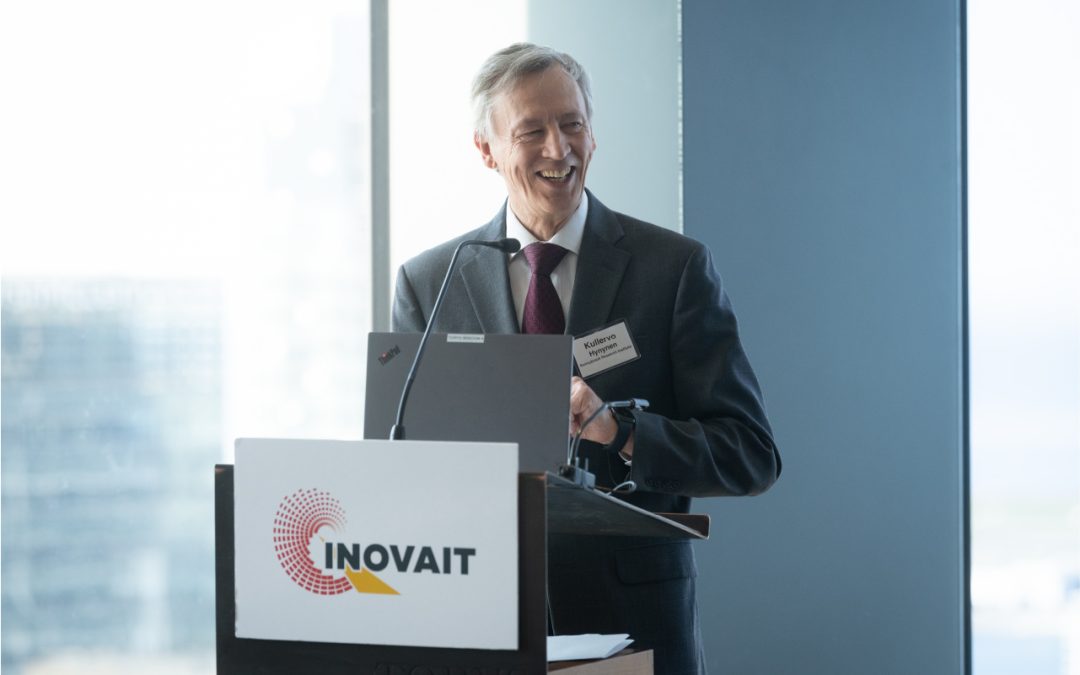 Dr. Kullervo Hynynen smiling and standing at a podium labeled INOVAIT in brightly lit and windowed conference room.