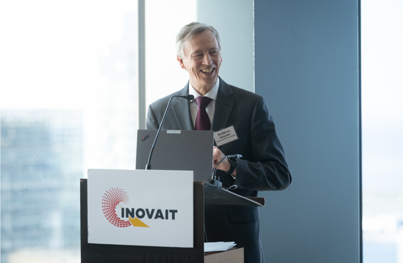Dr. Kullervo Hynynen smiling and standing at a podium labeled INOVAIT in brightly lit and windowed conference room.